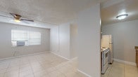 This single-floor unit offers a practical and straightforward layout, with light-toned ceramic tile flooring running consistently throughout the entire space. The open living and dining area is bright and airy, benefiting from multiple windows fitted with horizontal blinds that allow natural light to filter in. A ceiling fan with a built-in light fixture provides additional comfort and airflow in the main living space.

The kitchen is efficiently arranged in a galley-style configuration, featuring warm wood-toned cabinetry, granite-look laminate countertops, a stainless steel sink, a white electric range, and a full-size refrigerator. A peninsula counter provides a natural boundary between the kitchen and the adjacent dining or living area.

Multiple bedrooms are located along a central hallway, each equipped with window-mounted air conditioning units and windows that allow natural light and outdoor views. The neutral wall tones and tile flooring give each room a clean, consistent aesthetic.

A full bathroom is conveniently accessible from the hallway, featuring a white vanity with storage, a toilet, and a shower/tub combination with tiled surround. Built-in open shelving near the entryway adds useful storage. Textured ceilings are present throughout, complementing the unit's utilitarian yet comfortable character.