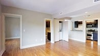 This single-floor residence presents a well-organized and functional layout with a clean, modern aesthetic. The main living area is finished with warm hardwood flooring that extends throughout, complementing the neutral gray walls and crisp white trim and baseboards. Recessed HVAC vents are integrated into the ceiling, contributing to a tidy, uncluttered appearance.

A standout feature of this unit is the in-unit laundry, conveniently housed behind a dedicated closet door and visible from the main living space, offering practical day-to-day convenience. Adjacent to the laundry area, a full bathroom is accessible directly from the main room, showcasing dark cabinetry that provides a rich contrast to the lighter walls and flooring.

Toward the opposite end of the space, a pass-through or open counter area connects to what appears to be a kitchen, where dark upper cabinetry is visible, maintaining a consistent design language throughout. The space benefits from a simple, open-plan configuration that allows for flexible furniture arrangement and a sense of airiness. Overall, the unit presents a practical and well-finished living environment with cohesive design elements and thoughtful use of space.