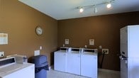 This shared laundry facility is a practical, well-organized space designed to serve its users with efficiency. The room is finished with warm, deep brown walls that create a grounded, neutral atmosphere, complemented by light tile flooring that is easy to maintain and keep clean. A row of top-loading commercial washing machines lines the wall, providing ample capacity for multiple users. Informational notices are posted above each machine, offering clear guidance on proper usage. Track lighting mounted on the white ceiling provides bright, even illumination throughout the space, ensuring visibility and comfort during use. A wall clock keeps users informed of the time, adding a thoughtful, practical touch to the room. A waste bin is conveniently positioned near the entrance for easy disposal. The overall layout is straightforward and uncluttered, making navigation simple and intuitive. This laundry room is a no-frills, utilitarian space that prioritizes function, cleanliness, and accessibility for all who use it.