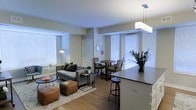 This single-floor unit showcases an open-concept layout that connects the kitchen, dining, and living areas into one cohesive and functional space. The kitchen features a sleek dark-topped island with bar seating, complemented by a modern rectangular pendant light that anchors the space. Hardwood-style flooring runs throughout, adding warmth and visual continuity across the entire floor plan.

The living area is comfortably proportioned, accommodating a tufted gray sofa, ottoman seating, and accent tables atop a patterned area rug. A floor lamp adds ambient lighting to the corner, enhancing the welcoming atmosphere. Adjacent to the living space, a compact dining area features a round wooden table with upholstered chairs, positioned near large windows fitted with white horizontal blinds that allow ample natural light to filter in.

The neutral color palette of soft whites and warm grays throughout creates a clean, modern aesthetic. The open layout promotes a sense of spaciousness, while thoughtful design details — from the pendant lighting to the well-placed furnishings — contribute to an overall ambiance that is both contemporary and inviting.