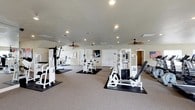 Located at 147 Links Dr in Canton, Mississippi, this dedicated fitness facility offers a well-designed, single-floor layout purpose-built for exercise and wellness. The space is generously sized, providing ample room for a full complement of professional-grade Cybex strength training machines, each positioned on protective rubber floor mats atop neutral carpeting. The open floor plan allows for comfortable movement between equipment stations without congestion. Recessed lighting is installed throughout the ceiling, ensuring bright, even illumination across the entire workout area. Ceiling fans are strategically placed to promote air circulation and comfort during exercise. The walls are painted in a clean, neutral palette, contributing to a bright and focused atmosphere. Decorative touches, including framed sports artwork and an American flag display, add character and motivation to the environment. Multiple interior doors suggest access to additional rooms or utility areas, and the overall layout reflects a thoughtful approach to functional fitness space design. This facility is well-suited for serious strength training and general fitness use.