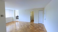 This single-floor space is characterized by its open and airy layout, highlighted by a distinctive parquet hardwood floor pattern that adds a classic, timeless charm to the interior. The living area is generously proportioned, offering ample room for a variety of furniture arrangements. Neutral white walls and standard-height ceilings contribute to a bright, clean atmosphere throughout.

A doorway to the left leads into a connected kitchen, which is visible from the main living area. The kitchen features what appears to be cabinetry, countertops, and standard appliances, and benefits from natural light through a window. The open sightline between the living area and kitchen creates a convenient flow for everyday living.

Toward the back of the living space, a central doorway leads to an additional room, likely a hallway or bedroom. To the right, a set of white bi-fold closet doors provides built-in storage. A secondary door on the far right appears to serve as an entry or exit point. The overall ambiance is understated and functional, offering a clean, move-in-ready canvas.