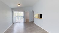 This single-floor space features an open-plan layout that maximizes its sense of spaciousness and functionality. The living area is finished with contemporary light-toned laminate flooring that runs throughout, complementing the soft, neutral wall color that contributes to the bright and airy atmosphere.

A key architectural feature is the sliding glass door fitted with vertical blinds, which opens onto a private balcony overlooking mature greenery. This connection to the outdoors allows abundant natural light to fill the interior, enhancing the overall sense of openness.

A pass-through window with a wooden countertop connects the living area to the kitchen, providing a practical and visually engaging design element. The kitchen area, visible through the pass-through, includes a sink and is illuminated by under-cabinet lighting, giving it a clean, functional aesthetic.

Crisp white baseboards frame the perimeter of the space, adding a polished finish. A flush-mount ceiling light provides additional illumination, and HVAC vents are integrated neatly into the walls. A doorway visible from the main living area suggests additional rooms beyond, completing this well-designed, move-in-ready space.
