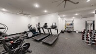 Located at 595 King St in Charleston, South Carolina, this fitness center offers a well-appointed exercise room designed to support a comprehensive workout experience. The space is finished with durable rubber flooring throughout, providing both safety and comfort underfoot, while crisp white walls and recessed lighting create a clean, bright atmosphere. Large ceiling fans are positioned throughout to promote air circulation during workouts.

The cardio zone features multiple treadmills and an elliptical trainer, each equipped with individual display screens, alongside a stationary bike to round out aerobic training options. A dedicated strength training area houses a cable machine, a squat rack with barbell and weight plates, and a flat bench, catering to a range of resistance training needs. A freestanding dumbbell rack stocked with a full set of weights offers additional versatility for functional and free-weight exercises.

Accessories including stability balls, resistance bands, and medicine balls are neatly organized on a storage rack, supporting flexibility and functional fitness routines. A dual water fountain station is set against a distinctive tiled accent wall featuring a bold pattern of teal and mauve tiles, adding a touch of character to the space. A glass-paned door provides access to the room, contributing to an open, welcoming feel throughout this well-organized fitness facility.