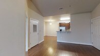 This single-story space showcases an open-concept design that seamlessly connects the main living area to the kitchen. The living space is distinguished by soaring vaulted ceilings that create an airy, expansive feel, complemented by warm hardwood flooring that extends throughout. Neutral-toned walls enhance the brightness of the interior, contributing to a clean and welcoming ambiance.

The kitchen features rich, dark wood cabinetry paired with a raised breakfast bar counter, offering both functional prep space and casual seating. Stainless steel appliances, including a refrigerator and microwave, are integrated into the cabinetry. A pendant light fixture above the kitchen area adds a focused accent, while a ceiling-mounted HVAC vent ensures year-round comfort.

A doorway near the entry leads to an adjacent room or exterior access, providing a logical flow through the space. Baseboards and door trim are finished in crisp white throughout, adding a polished contrast to the warm flooring. Overall, the layout balances openness with practical functionality, making it a versatile and comfortable living environment.