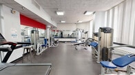 Located at 1320 W Somerville Ave in Philadelphia, PA, this fitness center offers a thoughtfully organized exercise space designed to support a full range of workout routines. The facility is laid out across a single open floor, providing a generous and unobstructed training environment.

The cardio zone features multiple treadmills, elliptical machines, a stationary air bike, and a rowing-style machine, all positioned near large windows fitted with vertical blinds that allow natural light to fill the space. The bright, airy atmosphere is further enhanced by recessed panel lighting set into an acoustic tile ceiling.

The strength training area is well-appointed with a variety of cable and resistance machines, a dumbbell rack with a full set of free weights, adjustable benches, and medicine ball storage. Full-length wall mirrors line one section of the gym, amplifying the sense of openness and allowing users to monitor their form during workouts. Bold red accent walls provide visual contrast against the clean white walls and dark rubberized flooring, contributing to an energetic and motivating ambiance.

A glass-partitioned section is visible along one wall, suggesting a separate area for administrative or supplementary use. The overall design is clean, functional, and professional, making this fitness center a comprehensive facility suited for a wide variety of training needs.