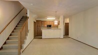 This two-floor home offers a well-organized and functional layout designed for comfortable everyday living. The main level showcases an open-concept design where the kitchen flows naturally into the living area, creating a connected and social atmosphere. The kitchen is appointed with warm honey-toned wood cabinetry offering generous storage, a built-in microwave, and a peninsula breakfast bar that serves as both a workspace and a casual dining spot. An elegant chandelier hangs above the peninsula, adding a decorative touch to the space. The living area is finished with neutral wall-to-wall carpeting that extends throughout the main floor, providing a warm and inviting feel underfoot. A staircase with a classic wooden banister leads to the upper level, adding architectural character to the open layout. The home's neutral color palette of soft whites and warm beiges lends itself to a clean and versatile aesthetic. Additional doorways visible from the main living area suggest further rooms beyond, hinting at a thoughtfully planned floor plan. The upper level expands the living space further, making this home a practical and welcoming residence suited for a variety of lifestyles.