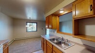 This single-floor unit offers a practical, well-organized layout with distinct living, kitchen, dining, and sleeping areas. The kitchen is designed as a galley-style space lined on both sides with warm oak cabinetry, offering ample upper and lower storage. White appliances, including a refrigerator, dishwasher, and electric range, are included. A double-basin stainless steel sink sits beneath upper cabinets, and tile flooring runs throughout the kitchen. A pass-through window connects the kitchen to the dining area, enhancing the open feel between spaces.

The dining area transitions seamlessly into the living room, which features soft carpeting and a chandelier pendant light overhead. A sliding glass door in the living area provides access to an outdoor space and allows natural light to fill the room. Baseboard heating units are present throughout, supplemented by a wall-mounted air conditioning unit in the bedroom.

The carpeted bedroom includes a spacious walk-in closet with built-in shelving and hanging rods, offering generous storage. A wood-framed window allows natural light and outdoor views into the room. The full bathroom features a single-sink vanity with a white countertop, a large mirror, tile flooring, and a full tub and shower combo.