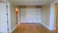 Located at 615 James St in Syracuse, New York, this single-floor space showcases a blend of classic architectural character and functional design. The main living area immediately draws attention with its striking, ornate white fireplace serving as a bold focal point. Flanking the fireplace are symmetrical built-in bookshelves, offering generous display and storage space while reinforcing the room's traditional aesthetic. Crown molding runs along the upper perimeter of the walls, adding an elegant finishing touch that speaks to the craftsmanship of the original construction. Warm-toned hardwood floors extend throughout the space, contributing a sense of continuity and timeless appeal. The neutral, earthy wall tones complement the white millwork, creating a balanced and inviting atmosphere. A glimpse into the adjacent area reveals a kitchen equipped with modern stainless steel appliances, indicating a thoughtful integration of contemporary convenience within the classic setting. The overall ambiance of the space is one of understated elegance — a property that honors its architectural heritage while remaining ready for modern use.