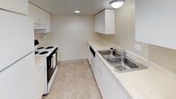 This single-floor unit is thoughtfully laid out across a elongated floor plan, offering a series of well-defined rooms finished in warm, neutral tones throughout. The living areas are generously proportioned, featuring soft wall-to-wall carpeting, crisp white baseboards, and crown molding that adds a classic architectural detail to the ceilings. Multiple large windows fitted with horizontal blinds allow abundant natural light to flow into each room, creating a consistently bright and open atmosphere.

The kitchen is efficiently designed with a galley-style layout, featuring white cabinetry, light countertops, a double stainless steel sink, and a full complement of appliances including an electric range and dishwasher. Light tile flooring anchors the space and extends into the bathrooms.

Two bathrooms serve the unit. One features classic hexagonal penny tile flooring, a pedestal sink, and a mirrored medicine cabinet—lending it a timeless character. The second bathroom is finished with larger format tile, a full tub-shower combination, and an extended vanity with ample storage.

The bedrooms are spacious and carpeted, each offering natural light through well-positioned windows. Crown molding appears in select rooms, reinforcing the unit's traditional character. Overall, the space presents a clean, neutral palette well-suited for a variety of uses.