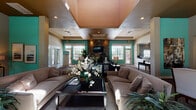 Located at 2120 Village Park Rd in Plant City, Florida, this single-story community clubhouse offers a thoughtfully designed gathering space that blends comfort with elegant architectural detail. The main lounge area immediately draws attention with its dramatic tray ceilings, recessed lighting, and bold teal accent walls that create a lively yet sophisticated atmosphere. A floor-to-ceiling stone fireplace serves as a striking focal point, complemented by a wall-mounted television and flanked by multi-pane windows that invite abundant natural light throughout the space. Comfortable neutral-toned sofas and upholstered seating are arranged around dark wood coffee tables, providing an inviting environment for socializing and relaxation. Lush tropical greenery and floral arrangements are positioned throughout, enhancing the warm, welcoming ambiance. Hardwood flooring in the entry areas transitions into plush carpeting across the lounge and office zones. A small refreshment bar, featuring decorative patterned wallcovering, granite countertops, and a coffered ceiling, offers added convenience near the entry. A dedicated office area with built-in workstations and executive seating provides a functional workspace. French doors open to views of the outdoor pool area, seamlessly connecting the interior to the surrounding Florida landscape.