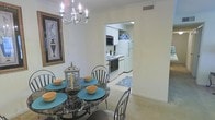 This single-floor residence offers a thoughtfully designed layout that connects living and functional spaces with ease. The dining area is an inviting focal point, highlighted by an elegant chandelier with candle-style lighting and crown molding that adds a refined architectural touch. A round marble-top dining table, surrounded by classic Windsor-style chairs, anchors the space and creates an intimate setting for meals and gatherings. Soft, neutral wall tones and plush carpeting contribute to the warm, comfortable ambiance throughout.

Adjacent to the dining area, the kitchen is accessible through an open archway, allowing for seamless flow between spaces. The kitchen features white cabinetry providing ample storage, a full-size refrigerator, a range with oven, and a built-in microwave. A decorative blue-and-white tiled accent wall adds a distinctive visual element, infusing the kitchen with personality and charm. The light-colored flooring in the kitchen contrasts pleasantly with the carpeted dining area, clearly delineating the two spaces while maintaining a cohesive overall aesthetic. This residence balances practicality with tasteful design details throughout.