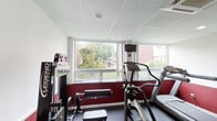 The Campus Edge fitness center in Carbondale, Illinois is a dedicated, single-floor exercise facility designed to support an active lifestyle. The room features a bold two-tone color scheme, with deep red accent walls on the lower half and crisp white above, creating an energetic and motivating atmosphere. Durable dark rubber flooring covers the entire space, providing a safe and practical surface for a variety of workouts.

The facility is equipped with a well-rounded selection of exercise equipment, including treadmills, an elliptical trainer, a cable weight machine, a multi-station strength machine, and free weights such as dumbbells. A wall-mounted television is positioned near the windows, allowing users to stay entertained during cardio sessions.

Large windows line one wall of the room, flooding the space with natural light and offering views of the surrounding greenery and campus exterior. A drop-panel ceiling with recessed lighting and a ceiling-mounted HVAC unit ensure year-round comfort.

A glass-panel door connects the fitness center directly to an adjacent common area, which features carpeted flooring, dining furniture, and wall décor including a large world map — making the transition between exercise and relaxation seamless. The overall layout is functional and efficiently organized to accommodate multiple users simultaneously.