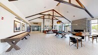 The Rock Pointe Clubroom in Rockaway, NJ is a thoughtfully designed two-floor amenity facility offering an impressive array of features across a generous footprint. The main level greets visitors with a soaring, cathedral-ceilinged lobby showcasing dramatic floor-to-ceiling windows, herringbone-patterned tile flooring, and an elegant stone accent wall framing a central fireplace and media display. Comfortable seating arrangements and curated artwork create a welcoming social atmosphere. Adjacent to the lobby, a game lounge with exposed beam ceilings features a billiards table, bar-height seating, and access to private conference and co-working rooms outfitted with warm wood furnishings, wall-mounted displays, and glass partition doors. A dedicated package and mailroom area offers modern smart lockers set against bold blue-accented walls. The expansive fitness center spans both floors and is the centerpiece of the facility, featuring a bright cardio zone lined with treadmills and cycling equipment alongside floor-to-ceiling windows overlooking lush greenery. The strength training area showcases a full complement of free weights, cable machines, and functional training rigs beneath high industrial ceilings with linear pendant lighting. A light-filled open staircase connects both levels, completing this comprehensive and polished amenity destination.