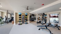 Located at 900 Hobbs Rd in Greensboro, NC, this modern fitness center is a comprehensively outfitted single-floor facility designed to support a wide range of workout routines. The space is organized into distinct functional zones, each dedicated to a specific training discipline, creating an efficient and intuitive layout for users of all fitness levels.

The cardio zone is positioned along a wall of large floor-to-ceiling windows, flooding the area with abundant natural light and offering views of the outdoor pool and surrounding community. This section houses multiple treadmills, elliptical trainers, stationary bikes, and a rowing machine, all complemented by ceiling fans that keep the environment comfortable.

The strength training area features a multi-station cable and weight machine alongside adjustable benches, with full-length mirrors lining the walls to assist with proper form. A dedicated free weights section includes a tiered dumbbell rack stocked with a comprehensive range of weights.

The functional training zone is equipped with medicine ball racks, stability balls, kettlebells, resistance bands, and foam rollers, all neatly organized on vertical storage towers. A lush vertical green wall adds a distinctive visual element to this section.

Throughout the facility, neutral tones, recessed lighting, and clean architectural lines contribute to a polished and professional ambiance. Dark barn-style entry doors provide a striking contrast to the light interior, completing the contemporary aesthetic of this well-appointed fitness center.