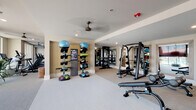 Located at 900 Hobbs Rd in Greensboro, NC, this modern fitness center is a comprehensively outfitted single-floor facility designed to support a wide range of workout routines. The space is organized into distinct functional zones, each dedicated to a specific training discipline, creating an efficient and intuitive layout for users of all fitness levels.

The cardio zone is positioned along a wall of large floor-to-ceiling windows, flooding the area with abundant natural light and offering views of the outdoor pool and surrounding community. This section houses multiple treadmills, elliptical trainers, stationary bikes, and a rowing machine, all complemented by ceiling fans that keep the environment comfortable.

The strength training area features a multi-station cable and weight machine alongside adjustable benches, with full-length mirrors lining the walls to assist with proper form. A dedicated free weights section includes a tiered dumbbell rack stocked with a comprehensive range of weights.

The functional training zone is equipped with medicine ball racks, stability balls, kettlebells, resistance bands, and foam rollers, all neatly organized on vertical storage towers. A lush vertical green wall adds a distinctive visual element to this section.

Throughout the facility, neutral tones, recessed lighting, and clean architectural lines contribute to a polished and professional ambiance. Dark barn-style entry doors provide a striking contrast to the light interior, completing the contemporary aesthetic of this well-appointed fitness center.