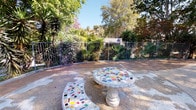 Located at Canyon in Oceanside, this outdoor courtyard offers a well-appointed communal gathering space designed to encourage relaxation and connection. The courtyard's most distinctive feature is its mosaic-tiled stone furniture — a round pedestal table and curved benches adorned with a vibrant patchwork of colorful tile fragments — providing a striking artistic focal point within the space.

The grounds are richly landscaped with a diverse mix of drought-tolerant and tropical plantings, including towering yucca trees, palms, flowering shrubs, and mature shade trees that frame the area and filter natural sunlight throughout the day. A wrought iron fence borders one side of the courtyard, beyond which additional greenery and hillside views extend the sense of openness.

A paved pathway winds through the courtyard, connecting the seating area to the surrounding multi-story buildings, which feature stucco exteriors, exterior staircases, and balconies in a Southwestern architectural style. A globe-style lamp post anchors the central walkway, adding a functional and decorative element. Patches of maintained lawn add softness between hardscaped surfaces. A secured gate provides access to an adjacent area, enhancing the courtyard's sense of enclosure while maintaining connectivity throughout the property.