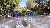Located at Canyon in Oceanside, this outdoor courtyard offers a well-appointed communal gathering space designed to encourage relaxation and connection. The courtyard's most distinctive feature is its mosaic-tiled stone furniture — a round pedestal table and curved benches adorned with a vibrant patchwork of colorful tile fragments — providing a striking artistic focal point within the space.

The grounds are richly landscaped with a diverse mix of drought-tolerant and tropical plantings, including towering yucca trees, palms, flowering shrubs, and mature shade trees that frame the area and filter natural sunlight throughout the day. A wrought iron fence borders one side of the courtyard, beyond which additional greenery and hillside views extend the sense of openness.

A paved pathway winds through the courtyard, connecting the seating area to the surrounding multi-story buildings, which feature stucco exteriors, exterior staircases, and balconies in a Southwestern architectural style. A globe-style lamp post anchors the central walkway, adding a functional and decorative element. Patches of maintained lawn add softness between hardscaped surfaces. A secured gate provides access to an adjacent area, enhancing the courtyard's sense of enclosure while maintaining connectivity throughout the property.