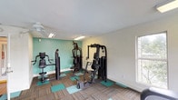 This fitness center is a thoughtfully designed, single-floor space dedicated to supporting a well-rounded workout routine. The room is immediately recognizable by its bold teal accent wall, which adds a vibrant, energizing touch to the otherwise neutral white walls and textured ceiling. The flooring features a distinctive checkerboard pattern of teal and striped carpet tiles, reinforcing the space's color scheme while providing a cushioned surface underfoot.

The strength training area is well-appointed with multiple cable and weight machines, including a multi-functional cable system and various resistance stations suited for a range of muscle groups. On the cardio side, the space accommodates treadmills and an elliptical trainer — including a Precor model — all positioned near large windows that frame views of surrounding greenery, allowing natural light to fill the room and making workouts feel less confined.

Overhead, a ceiling fan and fluorescent lighting fixtures ensure the space remains well-ventilated and properly illuminated. The room is accessible via a clearly marked exit door, and a hand sanitizer or paper towel dispenser is mounted on one wall for user convenience. Overall, this fitness center offers a functional, clean, and inviting environment for daily exercise.