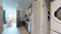 Micro Unit M2 is a single-floor living space expertly crafted to maximize functionality without sacrificing style. The open-concept layout makes intelligent use of the available footprint, seamlessly integrating the kitchen, living, and dining areas into one cohesive space. The kitchen is fully equipped with stainless steel appliances — including a refrigerator, range, microwave, and dishwasher — complemented by two-tone cabinetry featuring white upper cabinets and dark lower cabinets, quartz countertops, and a classic subway tile backsplash. Light hardwood flooring runs throughout, lending warmth and continuity to the space. The living area centers around a built-in Murphy bed system flanked by integrated storage cabinetry, allowing the room to transition effortlessly between a living and sleeping space. A compact dining area provides dedicated space for meals and entertaining. Open wall shelving adds a decorative and practical design element near the kitchen transition. The bathroom features tile flooring, a quartz-topped vanity with dark wood cabinetry, an illuminated mirror, and a walk-in tiled shower. Large windows allow natural light to brighten the interior, enhancing the overall sense of openness in this efficiently designed micro unit.