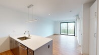 Located at 1237 N Van Buren St in Milwaukee's vibrant lower east side, this modern one-bedroom, one-bathroom unit offers a thoughtfully designed open-concept layout with clean lines and contemporary finishes throughout. Warm-toned hardwood flooring runs seamlessly across the entire space, complementing the crisp white walls and creating a bright, welcoming atmosphere.

The kitchen and living area flow together as one cohesive space. The kitchen features a large island with a white quartz countertop, an undermount sink, stainless steel appliances including a full-size refrigerator, built-in microwave, and range, plus dark cabinetry with sleek hardware. A linear pendant light anchors the kitchen island. The open living area benefits from a wall of floor-to-ceiling sliding glass doors that lead to a private balcony with city skyline views, filling the space with natural light.

The generously sized bedroom includes its own wall-mounted AC unit, a large black-framed window with treetop and skyline views, and direct access to a well-organized walk-in closet equipped with shelving and hanging rods. The bathroom is finished in all-white with a floating vanity, quartz countertop, large frameless mirror, and modern brushed-nickel fixtures.