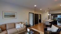 This single-floor space is designed with a clean, modern aesthetic that balances comfort and functionality. The living area serves as the heart of the unit, anchored by warm hardwood flooring that extends throughout, complemented by smooth, light-toned walls and recessed lighting that provides consistent, even illumination. A beige sofa with decorative throw pillows sits opposite a dark-toned coffee table, creating an inviting and well-composed seating arrangement. A glass-topped side table and a classic table lamp add warmth to the corner, while framed wall art contributes a finished, polished look to the space.

A standout architectural detail is the use of bold black-framed interior doors, which provide a striking contrast against the light walls and add a contemporary edge to the overall design. The doors lead to what appear to be a bedroom and a bathroom, visible in the background, suggesting a well-organized layout. A dining area with upholstered bar-style seating is positioned to the right, offering a functional transition between living and dining spaces. The consistent flooring, neutral color palette, and deliberate use of contrasting dark trim create a cohesive, stylish interior throughout.