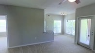This single-floor space is designed with an open, airy layout that promotes a sense of flow and connectivity throughout. The living area features soft, neutral-toned carpet and smooth gray walls that create a calm, understated backdrop. A ceiling fan overhead provides both function and subtle style, complementing the track lighting that illuminates the space with focused, adjustable light.

Large windows along the far wall flood the interior with generous natural light, offering views of the greenery outside and enhancing the overall sense of openness. The blinds on each window allow for flexible light control throughout the day.

A half-wall with a granite or stone-topped breakfast bar elegantly separates the living area from the kitchen, maintaining visual openness while defining each zone. This transitional element adds a modern, practical touch to the layout. The kitchen area, partially visible beyond the partition, benefits from the same abundant natural light.

The neutral color palette—soft grays and warm whites—carried throughout the space lends a clean, cohesive aesthetic that is both versatile and inviting. Vaulted or angled ceilings add architectural interest and contribute to the spacious feel of the interior, making this a well-appointed and thoughtfully designed living space.
