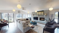 Located at 1944 Cambridge Ct in Palatine, Illinois, this single-floor community clubhouse offers a thoughtfully designed gathering space with distinct areas for relaxation, socializing, and fitness. The main lounge is an inviting, open space anchored by a floor-to-ceiling brick fireplace with a white mantel, providing a warm focal point. Vaulted ceilings, globe pendant lighting, a ceiling fan, and large windows along multiple walls flood the room with natural light and views of the surrounding outdoors. Wood-look flooring runs throughout, enhancing the cohesive, modern aesthetic. A passthrough window connects the lounge to adjacent spaces, maintaining an open, communicative flow. The dining area features a round marble-top table surrounded by plush navy velvet chairs, situated near a glass entry door that brings in additional natural light. The kitchen is outfitted with wood-toned cabinetry, white countertops, a textured tile backsplash, stainless steel appliances, and a breakfast bar with seating. The fitness center is divided across interconnected rooms, housing treadmills, an elliptical, a stationary bike, dumbbells, and a stability ball. Exposed ductwork adds an industrial character, while large windows and rubber flooring complete the functional space.