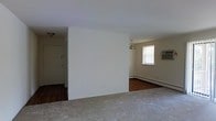 This single-floor space features an open layout divided into distinct zones by a central partial wall, creating a natural separation between areas while maintaining an open feel. The living area is finished with neutral-toned carpet, offering a soft and comfortable underfoot experience, while the adjacent room showcases hardwood flooring, adding warmth and contrast to the overall interior. The white walls throughout keep the space bright and versatile.

A window in the far room allows natural light to filter in, complemented by a window-mounted air conditioning unit for climate control. A baseboard heating unit runs along the wall, providing an additional source of warmth. A small chandelier with a classic design serves as a decorative focal point overhead in the adjoining room, adding a touch of character to the otherwise understated space.

Near the entrance, a door with a visible deadbolt provides secure access, and a ceiling-mounted light fixture illuminates the entryway. The layout flows naturally from the entry through the main living area, making the space functional and easy to navigate.