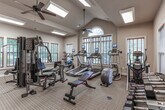 Located at 1650 S Fox Run Pkwy in Opelika, Alabama, this single-floor fitness and amenity facility offers a thoughtfully designed space catering to residents of all ages. The centerpiece is a generously sized fitness room featuring vaulted ceilings with a ceiling fan, an arched window, and multiple large windows dressed with wood-toned blinds that flood the space with natural light and views of the surrounding greenery. The gym is well-equipped with cardio machines including treadmills and ellipticals, a multi-station strength training unit, free weights with a dedicated dumbbell rack, medicine balls, and a flat workout bench, providing a comprehensive exercise experience. Towel service and posted fitness guidelines reflect a polished, community-oriented atmosphere. The facility connects via glass-paneled double doors to an interior corridor finished with stone-look tile flooring, leading seamlessly to additional areas within the building. A charming children's reading nook rounds out the amenity offerings, featuring a low table with child-sized seating, an assortment of picture books, playful toys, and vibrant pop-art prints on the walls — creating an engaging and welcoming corner for younger visitors. Together, these spaces combine functional fitness infrastructure with family-friendly touches in a warm, well-maintained setting.