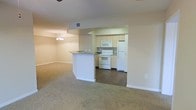 This single-floor unit features an open-concept layout that seamlessly connects the kitchen and living area. The kitchen is equipped with white appliances, including a freestanding electric range, over-the-range microwave, and a full-size refrigerator. Light-toned cabinetry lines the walls, providing ample storage, while a peninsula with a laminate countertop serves as a natural divider between the kitchen and the adjoining living space. The kitchen features dark hardwood-style flooring, which contrasts with the neutral-toned carpet that extends throughout the living area. The living space is generously proportioned, offering a comfortable, open feel. A chandelier adds a decorative touch above a designated dining area adjacent to the kitchen. The walls throughout are finished in a soft, neutral tone, contributing to a clean and understated aesthetic. Ceiling-mounted HVAC vents ensure year-round climate control. The overall ambiance is functional and straightforward, offering a practical layout suitable for everyday living.