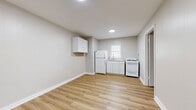 This efficiently designed 2-bedroom, 1-bathroom single-story home offers a practical and well-organized layout across its single floor. The interior is finished with light wood laminate flooring that runs consistently throughout all rooms, lending a cohesive and clean aesthetic to the space.

The living area is positioned near the front entry, which features a white door with an adjacent window, allowing natural light to brighten the room. The open kitchen area includes white shaker-style wall cabinets, a white refrigerator, a gas range, and a sink set beneath a window — providing functional essentials in a compact footprint.

The primary bedroom is a well-proportioned room with a window and direct access to a closet area with built-in shelving, which connects to the bathroom. The second bedroom is a smaller, bright room featuring a window and a wall-unit air conditioner. Both bedrooms are finished with the same warm wood flooring and neutral gray walls.

The bathroom is compact and efficient, featuring a pedestal sink, a medicine cabinet mirror, and a walk-in shower stall with chrome fixtures. Throughout the home, neutral gray walls, white trim, and recessed lighting contribute to a clean, minimalist feel that is functional and easy to personalize.