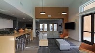 This contemporary single-floor space is thoughtfully designed to foster both casual interaction and focused gatherings. The open-plan layout is anchored by a spacious lounge area furnished with a modular gray sectional sofa, a sleek ottoman, and a vibrant orange accent chair, all arranged atop a patterned area rug. A large wall-mounted flat-screen TV serves as a focal point for the seating arrangement. Pendant lighting with cylindrical black fixtures hangs from the soaring ceilings, drawing the eye upward and lending a sense of dramatic height to the interior. A warm terracotta-toned accent wall adds depth and visual interest, contrasting beautifully with the surrounding neutral gray walls and light wood flooring. To one side, a bar-style counter with matching wood-tone stools provides an informal dining or working surface, complemented by decorative accents. Floor-to-ceiling glass doors and windows at the far end of the space flood the interior with natural light while offering views to the exterior beyond. A secondary area visible in the background suggests additional workspaces or meeting zones, making this a versatile and well-appointed environment suited for community use.