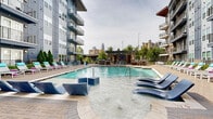 Located at 606 N Carolina Music Factory Blvd in Charlotte's dynamic NoDa district, this outdoor amenity space is designed for resort-style living with a variety of well-appointed recreational features. At the heart of the space is a generously sized swimming pool with a shallow sun shelf, where in-water chaise lounges invite residents to relax and soak in the atmosphere. The surrounding pool deck is finished with herringbone-pattern brick pavers and lined with colorful lounge chairs, creating a lively and welcoming environment. Framed by the contemporary multi-story building with modern grey paneling and private balconies, the space offers striking views of the Charlotte skyline. A dedicated lounge area features a large sectional sofa in vibrant teal with accent pillows, centered around a stone fire pit — ideal for evening gatherings. A pergola-covered outdoor kitchen is equipped with a stainless steel gas grill and prep stations, providing a fully functional space for outdoor cooking and dining. Lush greenery, including manicured topiaries and ornamental trees, adds natural contrast to the modern architectural backdrop. The cohesive design and thoughtful amenities make this outdoor space a standout communal destination in the heart of Charlotte.