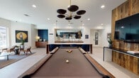 Located at 1104 E Belt Line Rd in Carrollton, Texas, this single-floor club room is a thoughtfully designed communal amenity space that balances entertainment and relaxation with a modern, upscale aesthetic. The open-concept layout flows seamlessly between distinct activity zones, all unified by large-format light tile flooring and crisp white ceilings with recessed lighting throughout.

At the heart of the space sits an expansive marble-topped island with dramatic veining, suspended above by sleek black-and-brass cylindrical pendant lights. The adjacent wet bar area is outfitted with warm wood cabinetry, a stainless steel sink, and appliances, making it ideal for hosting. A full-sized billiards table anchors the game area, illuminated by a bold multi-disc chandelier in black and gold, with a reflective tiled accent wall providing a striking visual backdrop.

The lounge zone features a dark mosaic tile feature wall framing a wall-mounted flat-screen television, complemented by leather armchairs and a sectional sofa atop a rich blue area rug. A separate seating nook benefits from abundant natural light streaming through tall windows with views of the outdoor grounds, furnished with mid-century modern chairs and curated artwork. A workspace area rounds out the room's versatile offerings, making this club room a dynamic hub for both socializing and productivity.