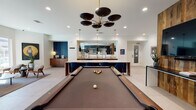 Located at 1104 E Belt Line Rd in Carrollton, Texas, this single-floor club room is a thoughtfully designed communal amenity space that balances entertainment and relaxation with a modern, upscale aesthetic. The open-concept layout flows seamlessly between distinct activity zones, all unified by large-format light tile flooring and crisp white ceilings with recessed lighting throughout.

At the heart of the space sits an expansive marble-topped island with dramatic veining, suspended above by sleek black-and-brass cylindrical pendant lights. The adjacent wet bar area is outfitted with warm wood cabinetry, a stainless steel sink, and appliances, making it ideal for hosting. A full-sized billiards table anchors the game area, illuminated by a bold multi-disc chandelier in black and gold, with a reflective tiled accent wall providing a striking visual backdrop.

The lounge zone features a dark mosaic tile feature wall framing a wall-mounted flat-screen television, complemented by leather armchairs and a sectional sofa atop a rich blue area rug. A separate seating nook benefits from abundant natural light streaming through tall windows with views of the outdoor grounds, furnished with mid-century modern chairs and curated artwork. A workspace area rounds out the room's versatile offerings, making this club room a dynamic hub for both socializing and productivity.