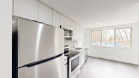 This thoughtfully designed 3-bedroom, 1-bathroom unit offers a clean, modern aesthetic across a single floor. The open kitchen is outfitted with full stainless steel appliances — including a refrigerator, electric range, microwave, and dishwasher — complemented by sleek white cabinetry that extends to the ceiling for ample storage. A large window floods the kitchen and adjacent living area with natural light, offering views of greenery outside. Light-toned hardwood-style flooring runs throughout the unit, creating a cohesive and airy feel.

The three bedrooms are well-proportioned, with the primary bedroom offering a comfortable footprint. Each room features crisp white walls, recessed lighting, and windows that bring in natural light and outdoor views. Black hardware accents on doors add a modern contrast to the otherwise all-white interior palette.

The hallways connect the spaces efficiently, maintaining a logical and functional flow. The full bathroom is finished in a clean white scheme, featuring a tiled tub-shower combination, a frosted window for privacy and natural light, and a vanity with a stone-topped countertop. The overall ambiance is bright, minimalist, and move-in ready, reflecting a fresh and contemporary design sensibility throughout.