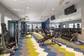 Located at 2001 S Hudson Ave in Boise, Idaho, this fitness center offers a well-appointed single-floor exercise room designed to support a wide range of workout routines. The space is generously sized, providing ample room for movement between equipment stations without feeling cramped.

The room features a striking chevron-patterned carpet in bold blue, yellow, and neutral tones, adding an energetic visual character to the space. Neutral gray walls and recessed ceiling lighting create a bright, clean atmosphere, while multiple windows fitted with blinds along the perimeter allow natural light to filter in throughout the day.

A full-length mirror along one wall visually expands the space and allows users to monitor their form during workouts. The equipment selection is comprehensive, including treadmills, elliptical trainers, and cycling machines for cardiovascular training, alongside a multi-station cable machine and weight bench for strength work. An extensive dumbbell rack offers a wide range of free weight options, and a TRX suspension training station is mounted near the windows. A BOSU balance trainer and additional accessories round out the functional fitness offerings.

Wall-mounted flat-screen televisions are positioned for viewing from multiple areas of the room. A ceiling fan provides additional air circulation. The space is accessed through a wood-paneled door and includes informational postings near the entrance, contributing to an organized and user-friendly environment.