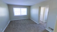This single-floor space presents a clean, straightforward layout designed for functional living. The featured room is finished with wall-to-wall neutral-toned carpeting that adds warmth underfoot, complemented by smooth, light gray-green painted walls and crisp white baseboards that frame the space neatly. A double-pane window along the exterior wall allows natural light to filter into the room, brightening the interior with a soft, even glow. The standard ceiling height and open wall space provide flexibility for various furniture arrangements. A doorway on the adjacent wall connects seamlessly to a neighboring room, suggesting a practical, flowing floor plan. The transition area between rooms features a clean threshold where the carpet meets what appears to be hard flooring in the adjoining space. Overall, the space maintains a neutral, move-in-ready aesthetic with a simple and unpretentious design character. The understated color palette and well-maintained finishes give the interior a tidy, welcoming atmosphere suited for everyday living.