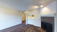 This single-floor space features an open-concept layout that connects the living area to the kitchen via a convenient pass-through window with a breakfast bar counter, allowing for easy interaction between the two spaces. The living room is a generous size, finished with dark wood-style flooring that adds warmth and contrast to the bright white walls. A standout feature is the tile-surround fireplace with a classic white mantel, positioned along one wall and serving as a natural focal point for the room. The kitchen, visible through the pass-through opening, includes wood-toned upper cabinetry and is well-lit, offering a functional workspace. A structural column divides the living and kitchen areas while preserving the open feel of the layout. The textured popcorn ceiling adds a traditional character to the space, and recessed ventilation and track lighting provide practical illumination throughout. A hallway visible from the main living area leads to additional rooms, suggesting a thoughtful floor plan that balances communal and private spaces. Overall, the unit presents a clean, neutral palette well-suited for comfortable everyday living.