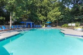 Located at 7843 Roswell Rd in Sandy Springs, Georgia, this outdoor recreational facility centers around a generously sized swimming pool with clear turquoise water, framed by a broad concrete pool deck ideal for lounging and socializing. The pool area is thoughtfully appointed with blue lounge chairs, chaise lounges, and shaded seating arrangements that provide both comfort and style.

An elevated wooden deck extends from the pool area, offering additional outdoor seating with round tables, chairs, and blue patio umbrellas that provide shade while overlooking the surrounding tree canopy. The verdant backdrop of mature trees creates a private, resort-like atmosphere throughout the space.

A dedicated pool house structure sits adjacent to the pool, providing a practical support facility for guests. Decorative white planters filled with colorful flowering plants and greenery add a welcoming touch to the transition between the deck and the pool deck below. The entire facility is enclosed by a fence, ensuring both safety and privacy. The combination of well-maintained landscaping, ample seating options, and thoughtful amenities makes this pool area a well-rounded outdoor retreat suited for both relaxation and recreation.