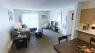 This single-floor space features an open layout that seamlessly connects the living and dining areas, creating a cohesive and functional environment. The living area is carpeted and generously sized, accommodating a sectional sofa, accent chair, and entertainment center with ease. Large windows dressed with vertical blinds allow abundant natural light to fill the room, contributing to a bright and welcoming atmosphere. A wall-mounted air conditioning unit provides climate control directly within the living space. The neutral wall tones in soft gray and white create a clean, versatile backdrop that complements a range of furnishing styles. A textured ceiling adds subtle dimension overhead. The entertainment area is anchored by a low-profile media console housing a flat-screen television, positioned conveniently across from the seating arrangement. Transitioning toward the dining area, a dark wood table and chairs are partially visible, suggesting a dedicated space for meals. The overall layout is practical and well-proportioned, with clear sightlines between zones that enhance the sense of openness throughout the interior.