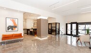 The lobby at 19 East, located at 20 E 19th St in Bayonne, NJ, presents a sophisticated and meticulously designed communal space that sets a refined tone for the building. The entrance area features expansive polished marble-style flooring that reflects the abundant ambient light, creating a bright and welcoming atmosphere. Floor-to-ceiling glass doors at the main entrance allow natural light to flood the interior while offering views of the surrounding streetscape.

A striking design feature of the lobby is the dramatic linear LED lighting system, with illuminated strips running across the ceiling and cascading vertically down accent walls, lending the space a distinctly contemporary aesthetic. The carefully curated décor includes bold abstract artwork, vibrant accent seating in warm orange tones, and tasteful architectural wall treatments that add depth and visual interest throughout.

The lobby seamlessly transitions into a well-appointed mail room, finished with dark herringbone-patterned wall tiles and a central wooden island counter, providing a functional yet stylish package and mail management area. Two elevators, framed by textured stone surrounds and polished tile flooring, offer convenient building access. The overall ambiance is polished and modern, reflecting a high standard of design and attention to detail throughout every shared area.