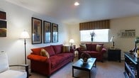 This single-floor space offers a comfortable and thoughtfully designed living environment. The living room serves as the centerpiece, featuring two plush red sofas arranged around a dark-finished rectangular coffee table, creating an inviting seating area ideal for relaxing or entertaining. Decorative throw pillows in warm, earthy tones complement the rich upholstery and add visual interest. The room is finished with light-colored carpet that softens the space and contributes to its welcoming ambiance. A set of three framed art pieces adorns one wall, adding a curated, polished touch to the interior. Natural light filters in through a window dressed with a patterned Roman shade in warm brown tones, balancing privacy with brightness. Recessed ceiling lighting provides clean, even illumination, while a classic table lamp adds a warm, ambient glow. A decorative floral arrangement and a sculptural centerpiece on the coffee table further enhance the room's refined aesthetic. The overall design combines functionality with a warm, residential character, making it a practical yet stylish living environment.