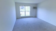 This single-floor space presents a straightforward and functional interior. The room shown is finished with neutral grey wall-to-wall carpeting, providing a soft underfoot feel and a clean, understated aesthetic throughout. White walls keep the space feeling bright and open, contributing to an airy atmosphere.

A large double-hung window serves as the room's focal point, allowing generous natural light to fill the space. The window frames a pleasant outdoor view of greenery, including grass and trees, which adds a sense of connection to the surrounding environment. A set of simple window treatments provides light control as needed.

Climate comfort is addressed through a wall-mounted air conditioning unit positioned above the window, as well as a baseboard heating unit running along the base of the exterior wall — offering year-round temperature regulation. The room's clean lines and empty layout make it highly versatile and adaptable to a variety of uses. Overall, this space offers a practical and tidy environment with key functional features already in place.