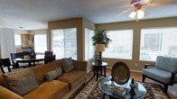 This single-story space centers around a well-appointed living room that immediately draws attention with its abundance of natural light. Large windows fitted with horizontal blinds line two walls, creating a bright corner effect that fills the room throughout the day. The warm, tan-toned walls complement the neutral aesthetic, while a textured ceiling with a ceiling fan overhead adds a classic residential character.

The living area is thoughtfully arranged, anchored by a plush, earth-toned sofa accented with decorative pillows. A patterned area rug defines the seating zone, beneath which sits a glass-topped coffee table styled with decorative accents. A small, dark round side table holds a traditional table lamp and a potted plant, adding organic warmth to the corner. Wood-look flooring runs throughout, providing a clean, durable foundation that ties the space together.

The overall ambiance is cozy yet functional, blending natural light with warm furnishings to create a welcoming atmosphere. The layout makes efficient use of the available space while maintaining an open, uncluttered feel suitable for comfortable daily living.
