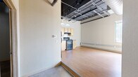 Located at 14 Fourth St E in downtown Saint Paul, Minnesota, this two-bedroom, one-bathroom loft-style residence offers a distinctive industrial character paired with an open, airy layout. The living and kitchen area is united by warm hardwood flooring and soaring ceilings with exposed structural beams, ductwork, and metal framework — hallmarks of converted urban loft architecture. Large windows along the perimeter flood the space with natural light and provide elevated panoramic views of the city and surrounding tree-lined landscape.

The kitchen features light maple cabinetry, dark countertops, black appliances including a range and refrigerator, and pendant lighting suspended from the exposed ceiling grid. The primary bedroom benefits from oversized windows offering sweeping outdoor views, while the second bedroom features concrete flooring and a structural column, adding to the loft's industrial aesthetic. The bathroom is functional and clean, with a wood-toned vanity, full tub-shower combo, polished concrete floors, and an accessibility grab bar. Throughout the unit, baseboard heating runs along the exterior walls, and neutral tones create a versatile, move-in-ready canvas in the heart of Saint Paul.