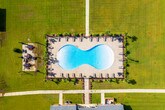 The outdoor amenity area centers around a large, freeform swimming pool filled with clear, blue water. The pool's curved shape adds a resort-like aesthetic, with depth markers visible along the edge indicating a gradual incline for varied swimmer comfort. The surrounding deck is finished in a warm, textured tan concrete, providing ample space for lounge seating and relaxation areas positioned around the perimeter.

Tall palm trees are strategically placed throughout the space, offering a tropical ambiance and partial shade. The pool area is enclosed by a black iron fence, providing both security and an open visual connection to the broader residential community beyond. Multiple lounge chairs and seating arrangements are distributed along the deck, accommodating a generous number of residents simultaneously.

In the background, multi-story residential buildings with yellow and tan exteriors and dark shuttered windows frame the space, contributing to an enclosed, community-centered feel. Additional landscaping, including low shrubs and ornamental plants, lines the fence perimeter, softening the hardscape. The wide-open sky above allows for abundant natural light, making this outdoor space bright and inviting throughout the day.