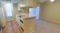 This single-floor space showcases an open-concept layout where the kitchen flows seamlessly into the living area. The kitchen is equipped with ample upper cabinetry in a cream finish, providing generous storage. A built-in dishwasher and a double-basin sink with a chrome faucet are integrated into the countertop, which extends outward to form a peninsula — a practical feature that doubles as a casual dining or workspace surface. Wood-look flooring runs throughout the kitchen, offering a contrast to the neutral carpeting that covers the living area. A window above the sink brings in natural light, brightening the kitchen workspace. The adjacent living area benefits from a sliding glass door fitted with vertical blinds, allowing for adjustable light control and potential outdoor access. Neutral wall tones throughout create a clean, cohesive backdrop. The textured ceiling adds a subtle architectural detail consistent across both spaces. Overall, the layout is functional and practical, designed to maximize the utility of a compact, single-level floor plan.