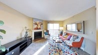 This single-floor residence offers a thoughtfully designed living area that blends comfort and personality in equal measure. The living room serves as the heart of the space, anchored by a classic white-mantled fireplace that adds warmth and architectural character. A bold, colorful piece of wall art depicting a longhorn cow adds an eclectic, artistic touch above the mantle, setting an expressive tone throughout the room.

Natural light flows generously into the space through a large sliding glass door, which leads to a private balcony overlooking a wooded outdoor setting — ideal for enjoying natural scenery through the seasons. A second window further enhances the bright, airy atmosphere of the room.

The living area is furnished with a striking terracotta-orange sofa accented with an assortment of decorative pillows in complementary tones, paired with a classic white armchair. A multi-colored geometric area rug anchors the seating arrangement, adding a lively, contemporary energy underfoot. A sleek media console along one wall provides practical storage, while a large decorative mirror reflects light and adds depth to the room. The warm yellow-toned walls and neutral ceiling tie the eclectic elements together into a cohesive and inviting living environment.